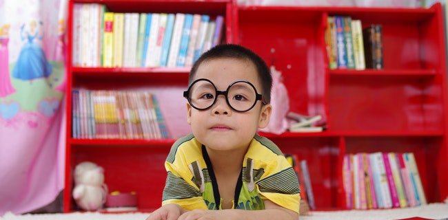 A toddler wearing glasses and reading a book next to the bookshelf