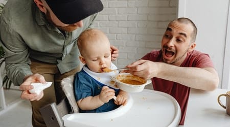 Men on a red T-shirt is feeding a baby on a high chair