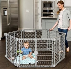 A baby is playing inside a baby gate and the mom is standing next to him